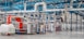 A spacious industrial facility with metal machinery and equipment, featuring a production line with a focus on textile processing. Brightly colored fabrics are moving through the machines, and there are large rolls of fabric and textile materials nearby. The environment is clean and organized, with a high ceiling and visible ductwork.