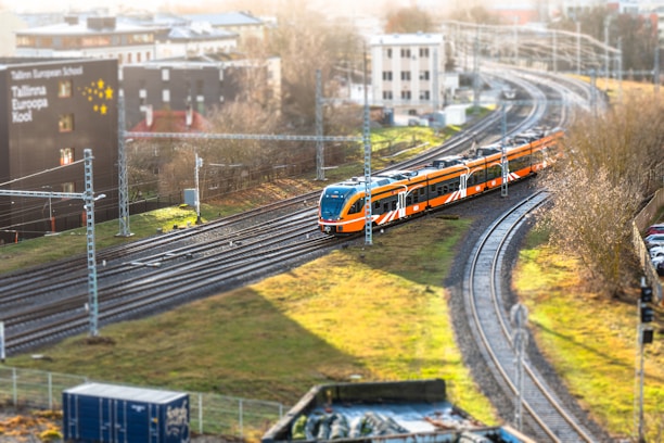 A vibrant orange and white train travels along a set of railway tracks, surrounded by grass and urban infrastructure. Buildings are visible in the background, and a sign for the Tallinn European School is prominent. The scene is lit by soft daylight, casting shadows on the ground.