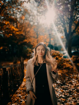 A stylish woman in her 40s wearing a soft beige trench coat, walking through a sunlit urban park with autumn leaves gently falling around her.