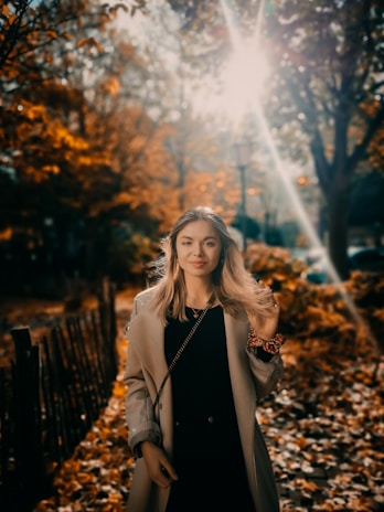 A woman wearing a light beige trench coat walking through a sunlit park with autumn leaves.