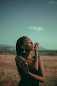 A woman stands in a serene landscape with her eyes closed, embracing a sense of tranquility. She wears a black dress and earrings, with her braided hair gently falling as sunlight casts a warm glow.