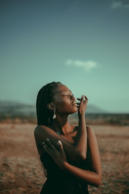 A woman stands in a serene landscape with her eyes closed, embracing a sense of tranquility. She wears a black dress and earrings, with her braided hair gently falling as sunlight casts a warm glow.