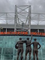 A group of three statues depicting football players stands in front of a modern stadium structure. The statues show the players with their arms around each other, with one holding a football. Above them is a large sign displaying the words 'Manchester United' in bold red letters. The background features a glass facade reflecting cloudy skies, with a white geometric metal framework on top.