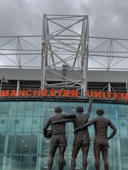 A group of three statues depicting football players stands in front of a modern stadium structure. The statues show the players with their arms around each other, with one holding a football. Above them is a large sign displaying the words 'Manchester United' in bold red letters. The background features a glass facade reflecting cloudy skies, with a white geometric metal framework on top.