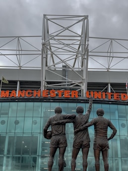 A group of three statues depicting football players stands in front of a modern stadium structure. The statues show the players with their arms around each other, with one holding a football. Above them is a large sign displaying the words 'Manchester United' in bold red letters. The background features a glass facade reflecting cloudy skies, with a white geometric metal framework on top.