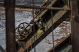 An industrial scene featuring rusted metal beams and machinery against a backdrop of a weathered brick wall. A large pulley system with visible chains is prominent, along with a sign indicating load capacity.