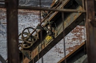 An industrial scene featuring rusted metal beams and machinery against a backdrop of a weathered brick wall. A large pulley system with visible chains is prominent, along with a sign indicating load capacity.