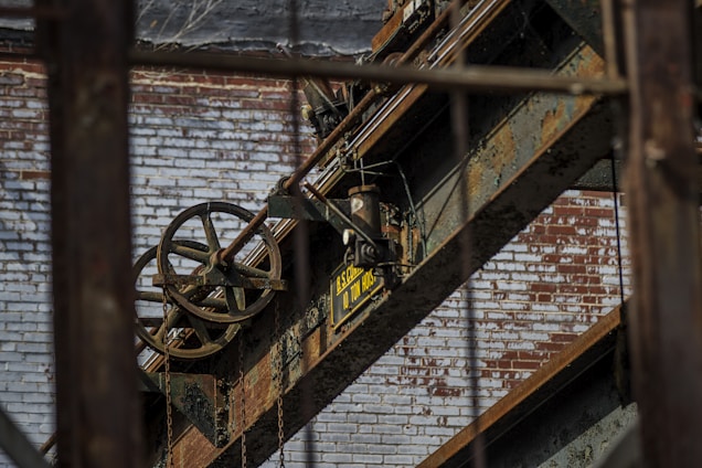An industrial scene featuring rusted metal beams and machinery against a backdrop of a weathered brick wall. A large pulley system with visible chains is prominent, along with a sign indicating load capacity.