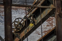 An industrial scene featuring rusted metal beams and machinery against a backdrop of a weathered brick wall. A large pulley system with visible chains is prominent, along with a sign indicating load capacity.