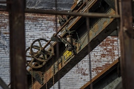 An industrial scene featuring rusted metal beams and machinery against a backdrop of a weathered brick wall. A large pulley system with visible chains is prominent, along with a sign indicating load capacity.