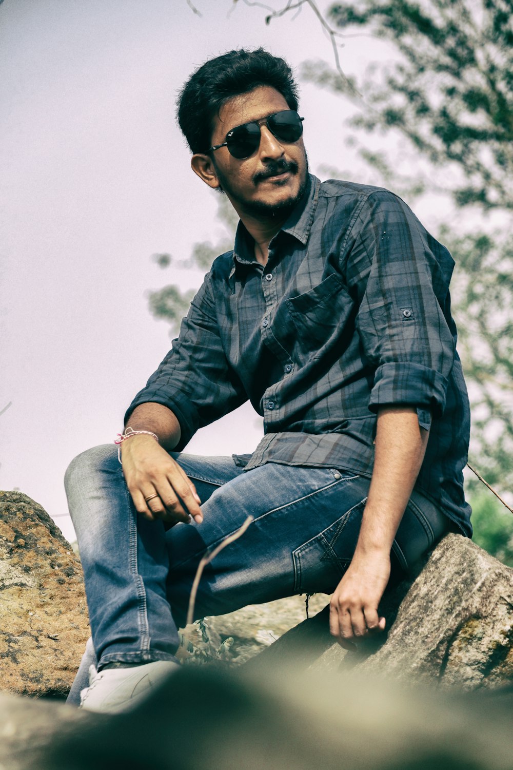 man in black and gray plaid button up shirt and blue denim jeans sitting on rock