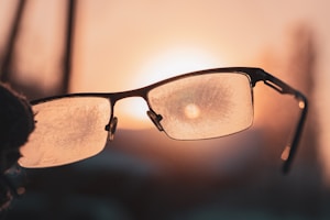 A pair of eyeglasses with frosted lenses is prominently featured against the backdrop of a warm, glowing sunset. The sunlight creates a halo effect through the frosty texture on the lenses, enhancing the ambient glow.