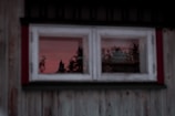 A wooden window frame with a reflection of a forest and red-tinted sky during sunset. The window is part of an old, weathered building with visible wood grain and peeling paint.