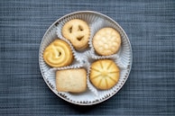 A round tin container holds five different types of cookies, each placed in its own white paper cup. The cookies include a pretzel shape, a circular one with a swirl, a round one with a stamped pattern, a rectangular sugar-topped cookie, and a circular star-patterned cookie. The background has a textured blue-gray fabric surface.