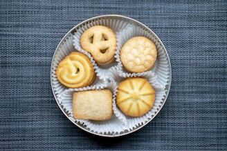 A round tin container holds five different types of cookies, each placed in its own white paper cup. The cookies include a pretzel shape, a circular one with a swirl, a round one with a stamped pattern, a rectangular sugar-topped cookie, and a circular star-patterned cookie. The background has a textured blue-gray fabric surface.