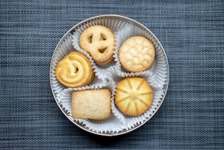 A round tin container holds five different types of cookies, each placed in its own white paper cup. The cookies include a pretzel shape, a circular one with a swirl, a round one with a stamped pattern, a rectangular sugar-topped cookie, and a circular star-patterned cookie. The background has a textured blue-gray fabric surface.