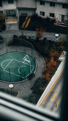 An aerial view of an outdoor basketball court surrounded by a mesh fence, situated in a residential area with neatly arranged pathways and landscaping. The scene includes autumnal trees and shrubs with vibrant colors and a portion of a road with a yellow directional arrow.