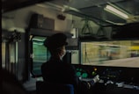 A train conductor is seated in the control cabin, wearing a uniform and mask. The view outside is blurred, suggesting motion. The cabin is lit with fluorescent lights, and various control panels and dials are visible.