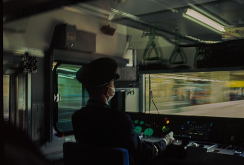 A train conductor is seated in the control cabin, wearing a uniform and mask. The view outside is blurred, suggesting motion. The cabin is lit with fluorescent lights, and various control panels and dials are visible.