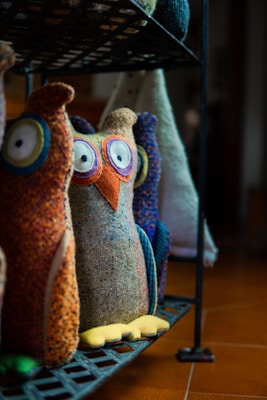 Colorful fabric owls with large eyes and various patterns are displayed on a dark metal shelf. The owls are crafted with different textured materials, featuring vibrant colors like orange, blue, and yellow. The background is softly blurred, emphasizing the playful and artistic design of these decorative pieces.