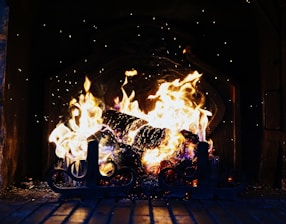 A roaring fire in a fireplace, with bright flames and glowing embers creating a warm and inviting atmosphere. Sparks occasionally rise from the fire, and a set of decorative metal fireplace tools can be seen in the foreground.