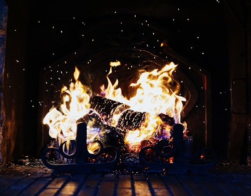 A roaring fire in a fireplace, with bright flames and glowing embers creating a warm and inviting atmosphere. Sparks occasionally rise from the fire, and a set of decorative metal fireplace tools can be seen in the foreground.