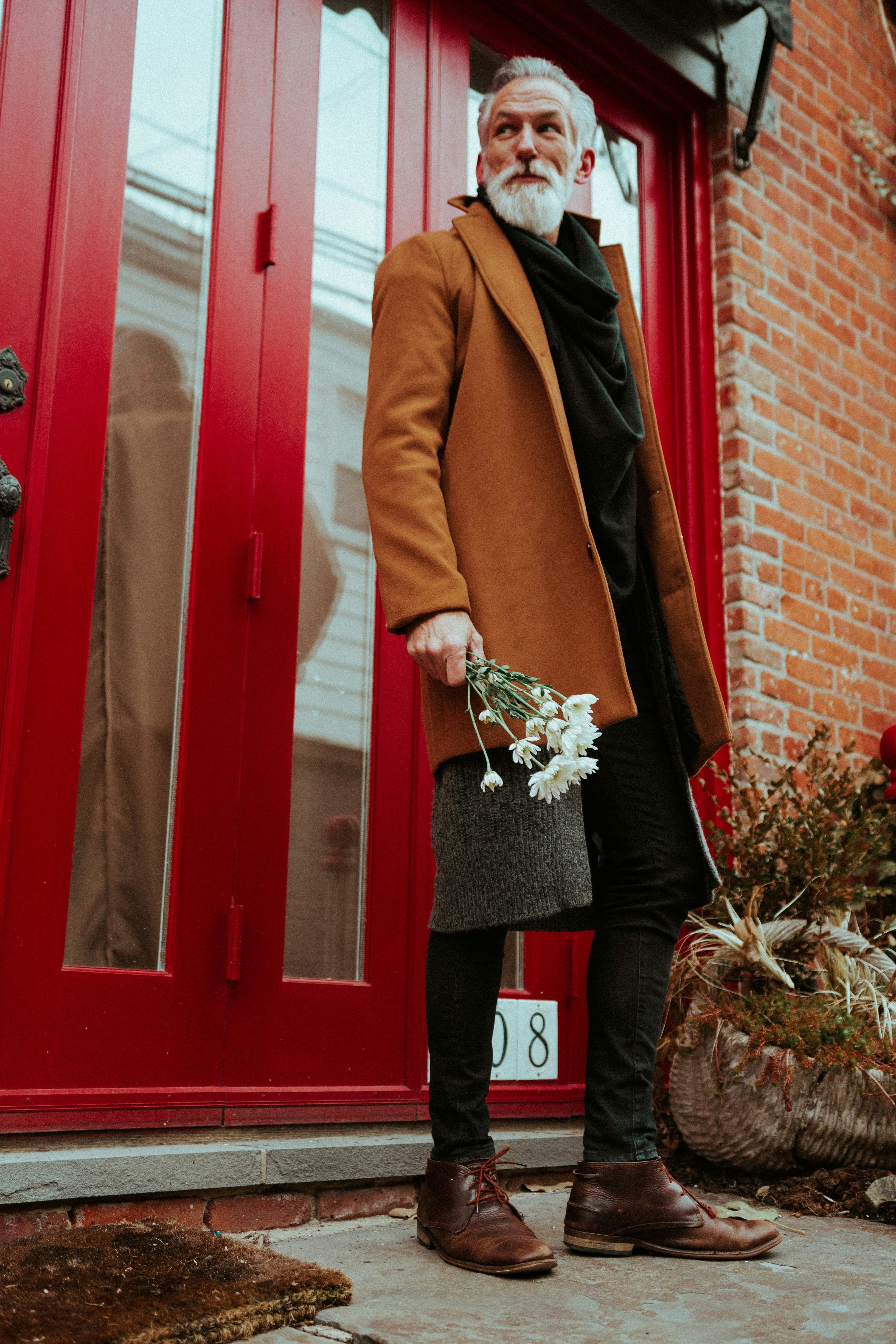 woman in brown coat holding bouquet of flowers standing beside red wooden door