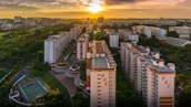 Aerial view of a residential area with several high-rise apartment buildings lined up. The backdrop is an expansive cityscape under a dramatic sunset sky, with vibrant orange and yellow hues. A sports court is visible in the foreground, surrounded by lush greenery and well-maintained public areas.
