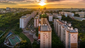 Aerial view of a residential area with several high-rise apartment buildings lined up. The backdrop is an expansive cityscape under a dramatic sunset sky, with vibrant orange and yellow hues. A sports court is visible in the foreground, surrounded by lush greenery and well-maintained public areas.