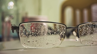 A close-up view of a pair of eyeglasses resting on a surface, with water droplets scattered across the lenses. The background appears blurred, showcasing bokeh effects with light reflections. The glasses have a metal frame and the surface seems smooth and light-colored.