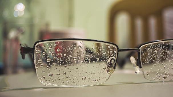 A close-up view of a pair of eyeglasses resting on a surface, with water droplets scattered across the lenses. The background appears blurred, showcasing bokeh effects with light reflections. The glasses have a metal frame and the surface seems smooth and light-colored.