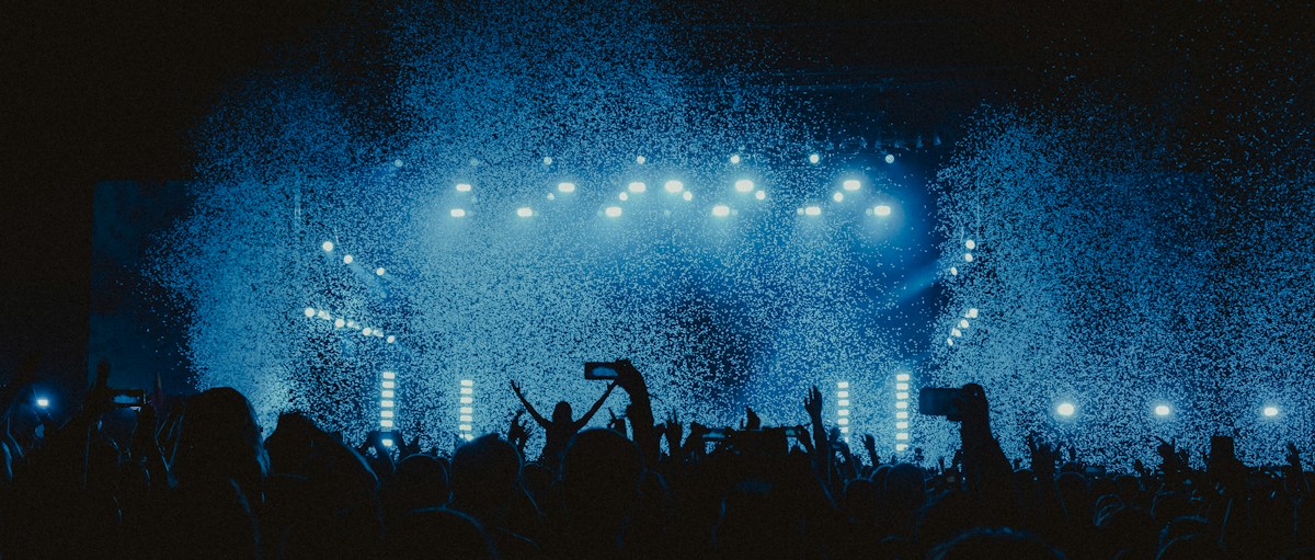 Une foule lève les bras et filme la scène d’un concert de nuit, sous des projecteurs bleus et une pluie de confettis