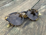 A pair of tortoiseshell sunglasses with round lenses rests on a weathered wooden surface. The lenses reflect palm trees and a clear sky, adding a natural touch to the scene.