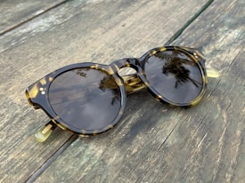 A pair of tortoiseshell sunglasses with round lenses rests on a weathered wooden surface. The lenses reflect palm trees and a clear sky, adding a natural touch to the scene.