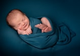 A newborn baby is swaddled in a teal fabric, sleeping peacefully on a matching teal background. The baby has a content smile on their face and their hands are gently tucked under their chin.