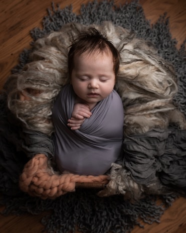 A peacefully sleeping baby wrapped in a soft, gray fabric, nestled in a cozy arrangement of textured, woolen materials. The baby has closed eyes and slightly tousled hair, with hands gently clasped. The setting includes soft layers of gray and beige hues, providing a warm and snug environment.