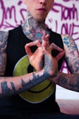 A tattooed individual is holding their hands in a mudra gesture. The person is wearing a black sleeveless shirt with a yellow smiley face design. Tattoos cover their arms, neck, and hand, including a skyline on the forearm. The background features purple graffiti-style writing.