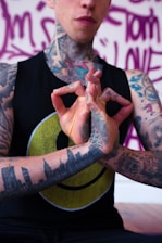 A tattooed individual is holding their hands in a mudra gesture. The person is wearing a black sleeveless shirt with a yellow smiley face design. Tattoos cover their arms, neck, and hand, including a skyline on the forearm. The background features purple graffiti-style writing.