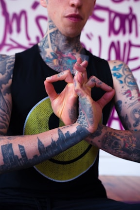 A tattooed individual is holding their hands in a mudra gesture. The person is wearing a black sleeveless shirt with a yellow smiley face design. Tattoos cover their arms, neck, and hand, including a skyline on the forearm. The background features purple graffiti-style writing.