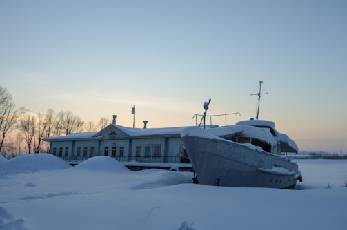 A snow-covered boat is docked beside a building that is partially obscured by snow drifts. The scene is set during early evening or late afternoon with the sky showing hues of pale orange and blue. Bare trees are visible in the background, indicative of winter.