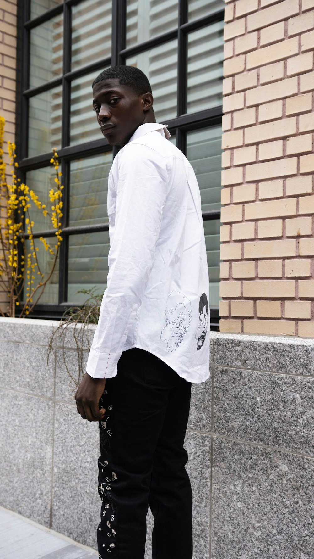 Hombre con blanca de manga larga y pantalón negro parado al lado de una pared de ladrillo marrón durante el día – Imagen Estilo gratis en Unsplash