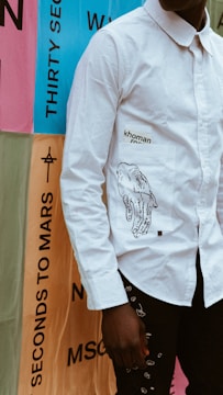 A person wearing a white long-sleeved shirt with an illustration of hands and text printed on the pocket. The background features colorful posters with large text, including phrases like 'THIRTY SECONDS TO MARS'. The scene suggests a contemporary or urban setting.