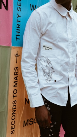 A person wearing a white long-sleeved shirt with an illustration of hands and text printed on the pocket. The background features colorful posters with large text, including phrases like 'THIRTY SECONDS TO MARS'. The scene suggests a contemporary or urban setting.
