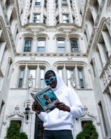 A person wearing a white hoodie and blue sunglasses is standing in front of an ornate, historic-style building. They are reading a Forbes magazine, which prominently features the word 'Billionaires' on the cover.