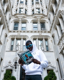 A person wearing a white hoodie and blue sunglasses is standing in front of an ornate, historic-style building. They are reading a Forbes magazine, which prominently features the word 'Billionaires' on the cover.