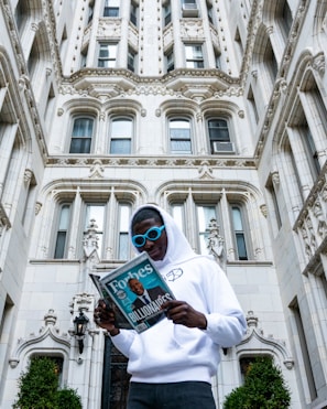 A person wearing a white hoodie and blue sunglasses is standing in front of an ornate, historic-style building. They are reading a Forbes magazine, which prominently features the word 'Billionaires' on the cover.