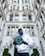 A person wearing a white hoodie and blue sunglasses is standing in front of an ornate, historic-style building. They are reading a Forbes magazine, which prominently features the word 'Billionaires' on the cover.