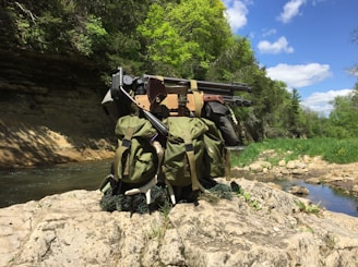 A tactical backpack with various equipment, including a sniper rifle, sits on a rocky ledge beside a stream. The setting is a lush, forested area with bright green trees under a blue, partially cloudy sky.