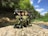 A tactical backpack with various equipment, including a sniper rifle, sits on a rocky ledge beside a stream. The setting is a lush, forested area with bright green trees under a blue, partially cloudy sky.