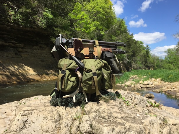 A tactical backpack with various equipment, including a sniper rifle, sits on a rocky ledge beside a stream. The setting is a lush, forested area with bright green trees under a blue, partially cloudy sky.